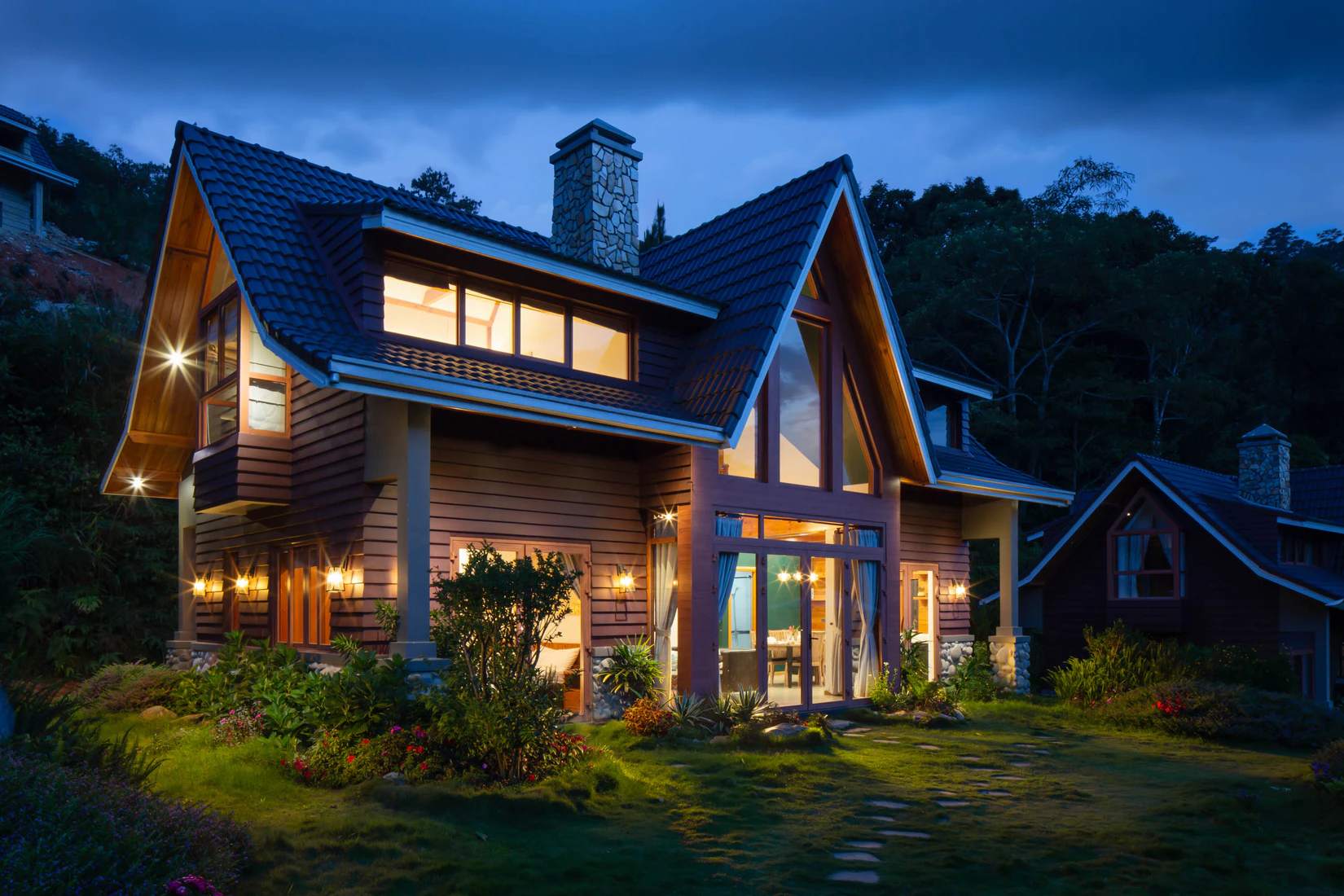 A wooden house with a garden and a chimney at nighttime with the lights on