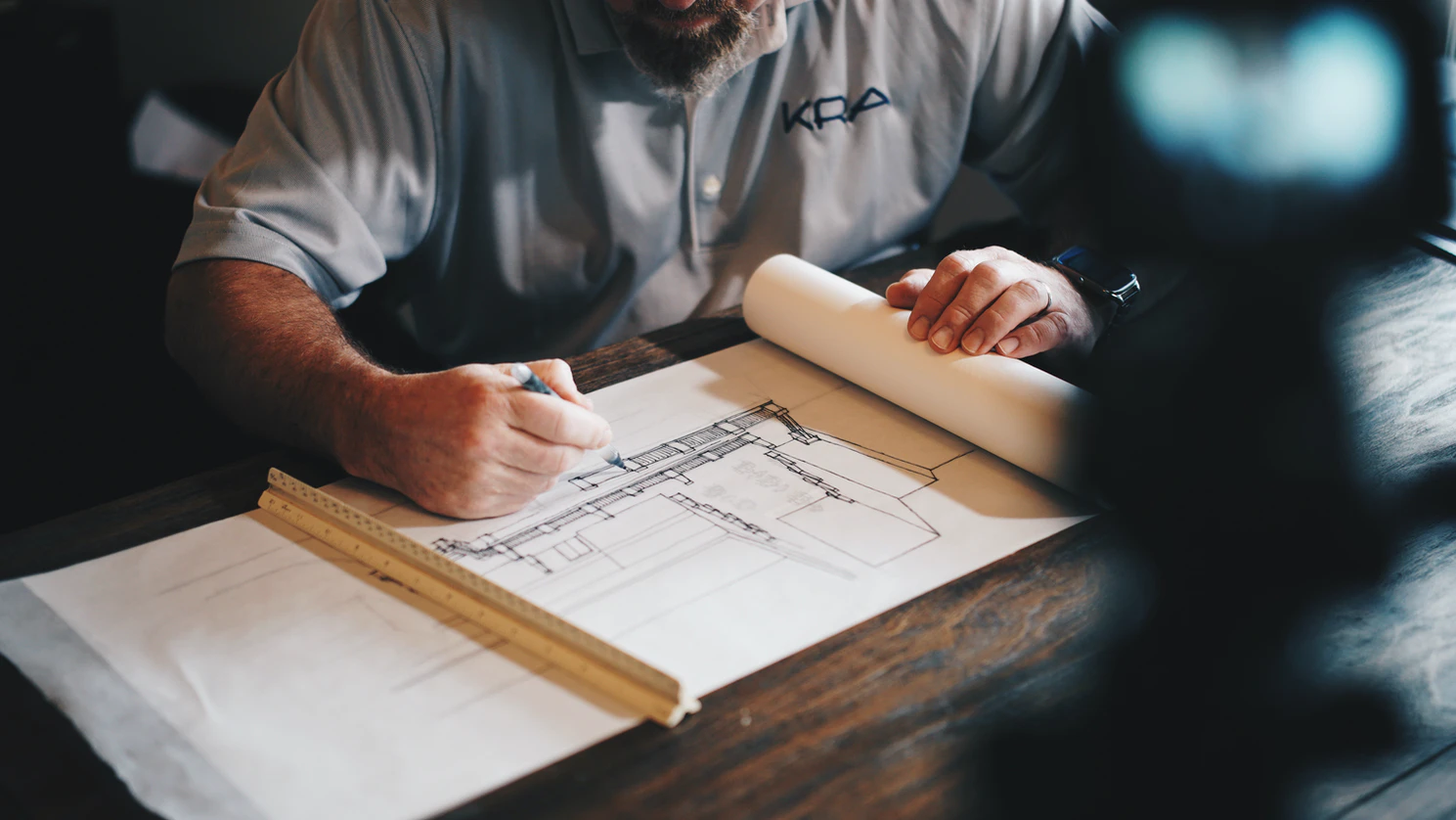 Man with a beard sitting at a wooden table with an architectural blueprint, pencil and a ruler