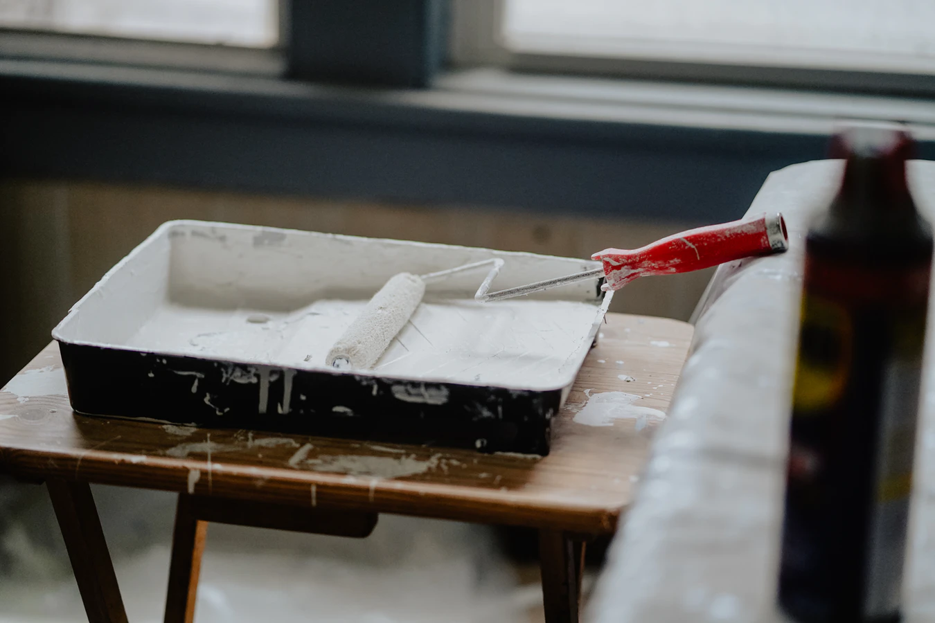 A bucket of white paint on a wooden table with a paint roller that has a red handle