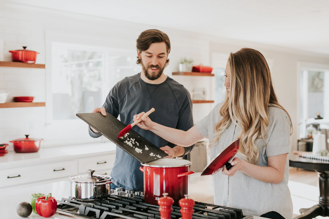 A man with a beard and a women with blonde hair cooking together in the kitchen with a red pot and black chopping board