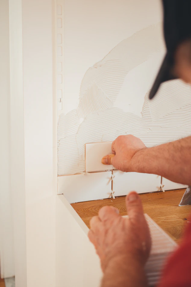 a man with a hat on applying groute and white ceramic tiles to a new wall